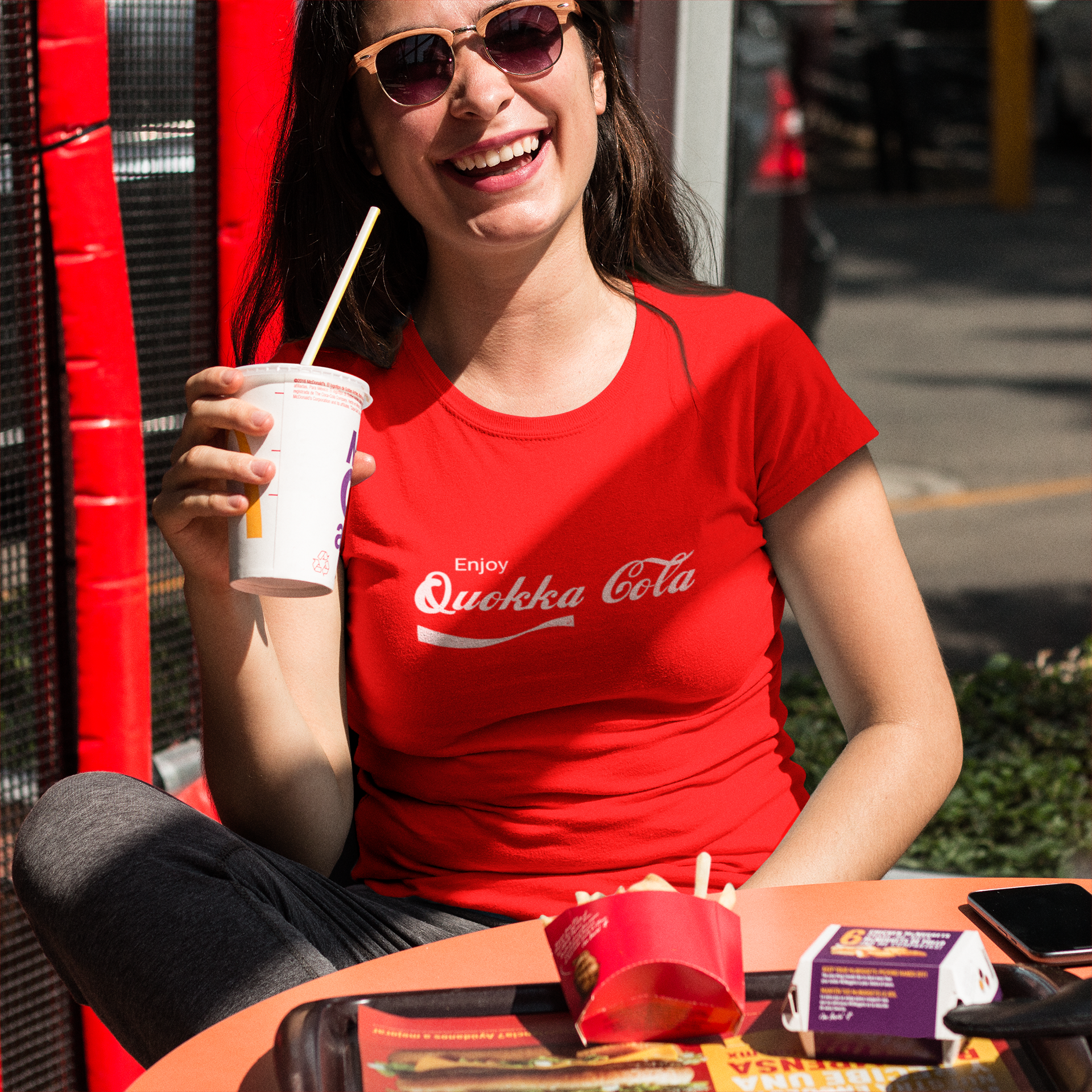 Smiling woman wearing a red 'Enjoy Quokka Cola' T-shirt, enjoying a drink outdoors at a casual fast-food setting. Fun and vibrant casual style.